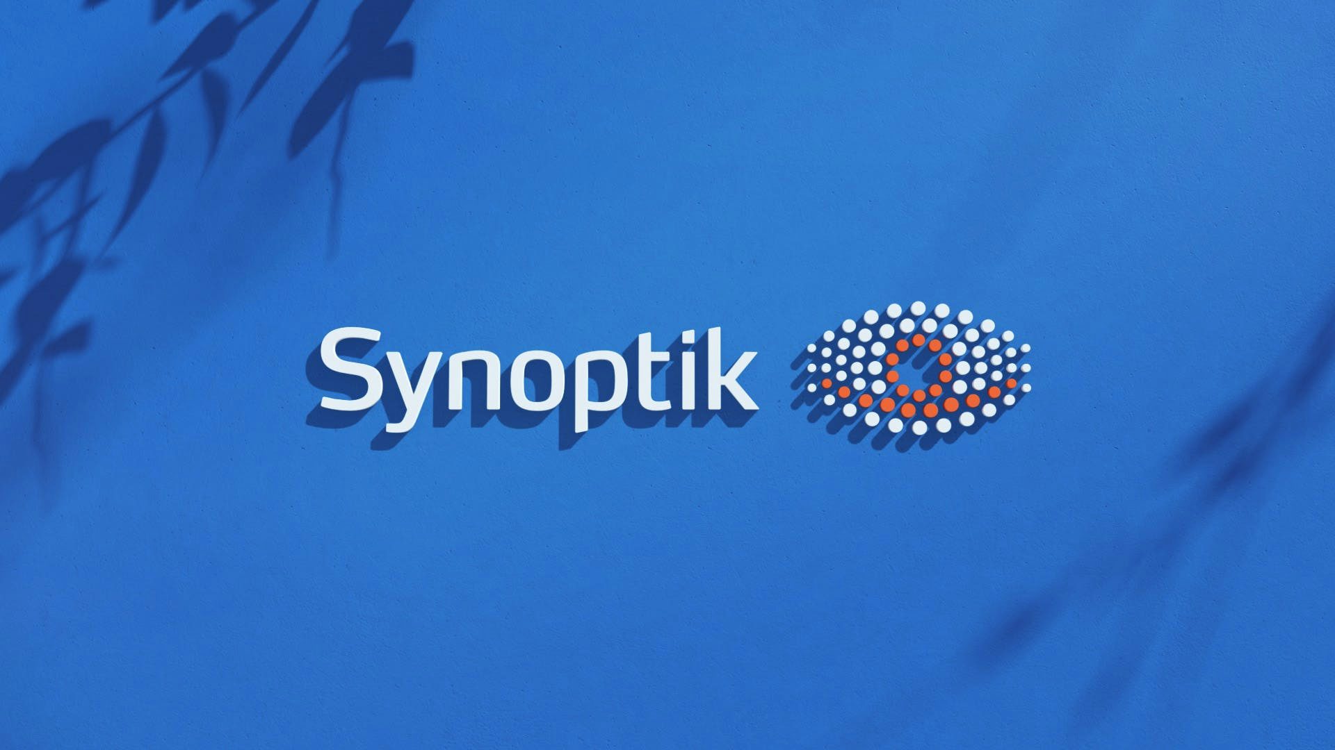 The synoptik logo extruded from a blue wall. Shadows of branches on the wall.